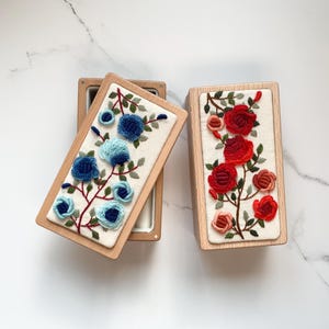 May include: Two rectangular wooden boxes with embroidered floral designs. One box features blue flowers, the other red. The embroidery is on a cream-colored background, framed by light wood. The boxes are open, revealing a white interior.