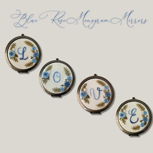 May include: Four small round compact mirrors with a white background and blue and gold embroidery. Each mirror has a different letter embroidered on it, spelling out the word "LOVE".