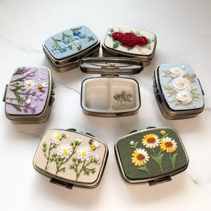 May include: A collection of small, rectangular metal boxes with hinged lids. Each box features embroidered floral designs in various colors, including blue, red, white, purple, and yellow. One box is open, revealing a compartment with jewelry.