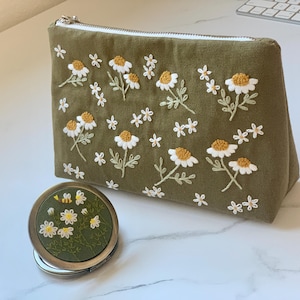 May include: Olive green embroidered cosmetic bag and matching compact mirror. The bag and mirror feature white daisies with yellow centers and green leaves. The bag has a silver zipper.