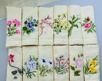 May include: A set of 15 off-white linen tea towels with embroidered floral designs. Each towel features a different floral motif, including roses, carnations, lilies, and daisies.