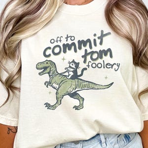 May include: A cream-coloured t-shirt with the text "off to commit tomfoolery". The graphic shows a green dinosaur with a black cat riding it. The shirt has short sleeves and a loose fit. The text and graphic are in shades of green and black.