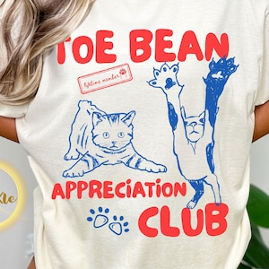 May include: Ivory Comfort Colors T-shirt with a blue and red graphic. The design features two cats, the text "TOE BEAN APPRECIATION CLUB", and a "lifetime member" stamp. The shirt is made of 100% cotton.
