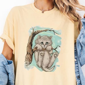 以下が含まれることがあります： 木にぶら下がっている灰色の猫のイラストが描かれた、明るい黄色のTシャツ。猫は大きな緑色の目を持ち、水色の背景に配置されています。Tシャツは半袖で、ゆったりとしたフィット感です。