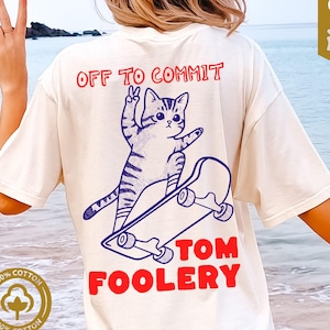 May include: Ivory Comfort Colors T-shirt with a graphic of a cat on a skateboard, the text "OFF TO COMMIT" above the cat, and "TOM FOOLERY" below. The shirt is 100% cotton.