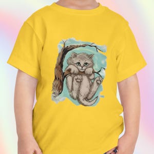May include: Yellow short-sleeved t-shirt featuring a cartoon illustration of a gray kitten hanging from a tree branch. The kitten has a cute expression, and the background is light blue with white accents.