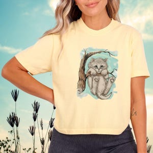 May include: A light yellow t-shirt featuring a gray kitten hanging from a tree branch. The kitten has big eyes and is set against a light blue background. The shirt is short-sleeved and has a crew neck.
