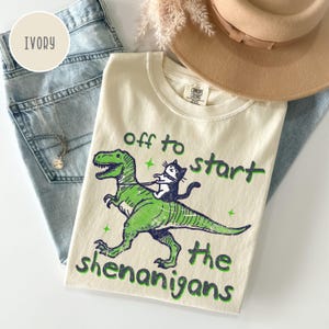 Può includere: T-shirt Comfort Colors color avorio con grafica di dinosauro verde e gatto bianco e nero. Il testo "off to start the shenanigans" è verde.