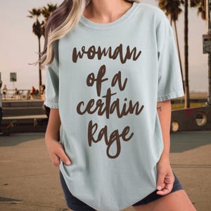 May include: A light blue t-shirt with the text "woman of a certain rage" in brown script. The shirt has short sleeves and a relaxed fit. The background shows a beach scene with palm trees.