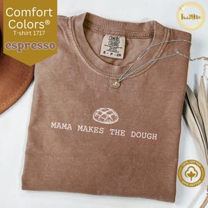 Può includere: Una maglietta marrone con il testo "Mama Makes The Dough" e un disegno di un pane. La maglietta è realizzata in 100% cotone.