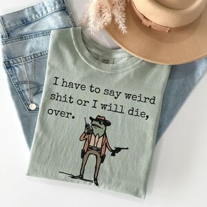 Puede incluir: Camiseta verde claro con el texto "I have to say weird shit or I will die, over." y una rana de dibujos animados con sombrero y pistola. La camiseta se combina con vaqueros azules y un sombrero beige.