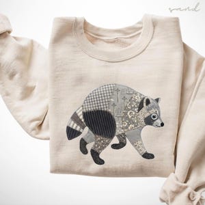 May include: A sand-colored crewneck sweatshirt featuring a large, central graphic of a raccoon. The raccoon is composed of various gray and white fabric patterns, including gingham, stripes, and floral prints, with a faint outline of the Seattle skyline.