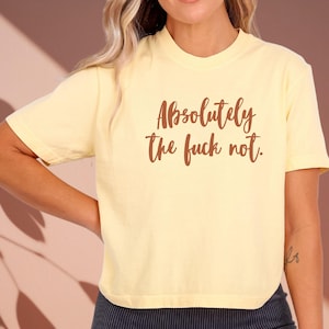 May include: A light yellow t-shirt with the phrase "Absolutely the fuck not" printed in brown script. The shirt has short sleeves and a crew neck. The image is a close-up of the shirt.