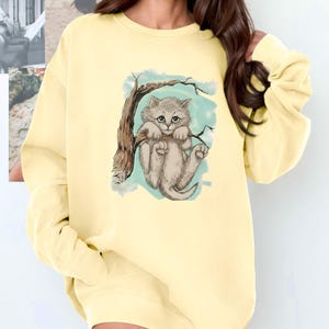 May include: A pale yellow sweatshirt featuring a whimsical illustration of a gray kitten clinging to a tree branch. The artwork is set against a light blue backdrop, creating a playful and charming design. The sweatshirt has long sleeves and a crew neck.