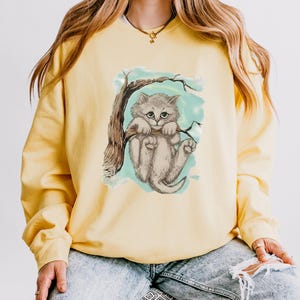 Pode incluir: Camisola amarela com uma ilustração de um gatinho cinzento pendurado num galho. O gatinho tem olhos verdes e está sobre um fundo azul claro. A camisola é de cor amarela lisa.
