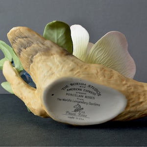 May include: A porcelain Peace Rose figurine by The Boehm Studios and American Express. The figurine is a white rose with green leaves and a pink petal. The base is a brown, textured piece of wood. The bottom of the base has the text "The Boehm Studios and American Express present Porcelain Roses from The World's Legendary Gardens Peace Rose made in USA."