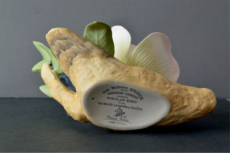 May include: A porcelain Peace Rose figurine by The Boehm Studios and American Express. The figurine is a white rose with green leaves and a pink petal. The base is a brown, textured piece of wood. The bottom of the base has the text "The Boehm Studios and American Express present Porcelain Roses from The World's Legendary Gardens Peace Rose made in USA."