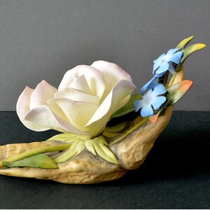 May include: A porcelain figurine of a white rose with pink edges, nestled in a hand-shaped base with green leaves and blue forget-me-not flowers.