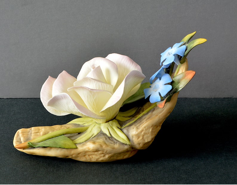 May include: A porcelain figurine of a white rose with pink edges, nestled in a hand-shaped base with green leaves and blue forget-me-not flowers.