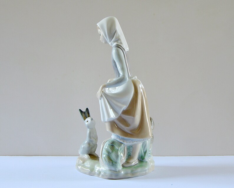 May include: A porcelain figurine of a girl in a blue and white dress and a white bonnet, holding a small white rabbit. The figurine is standing on a base with green and brown foliage.