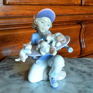 May include: A porcelain figurine of a child wearing a blue shirt and a purple baseball cap holding a skateboard with four kittens on it.