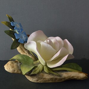 May include: A porcelain figurine of a white rose with pink edges, surrounded by green leaves and blue flowers, resting on a brown, textured base.
