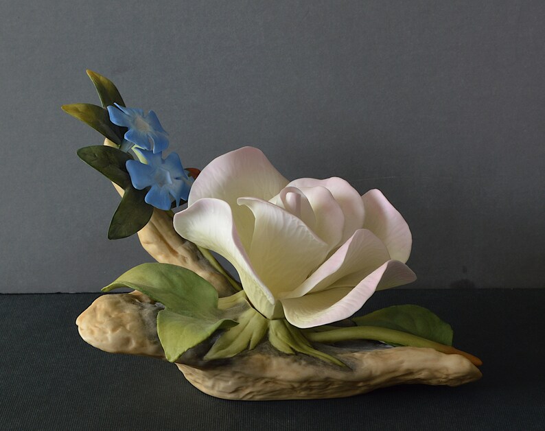May include: A porcelain figurine of a white rose with pink edges, surrounded by green leaves and blue flowers, resting on a brown, textured base.