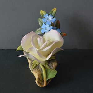 May include: A porcelain figurine of a white rose with pink highlights and blue flowers. The rose is on a brown base with green leaves.