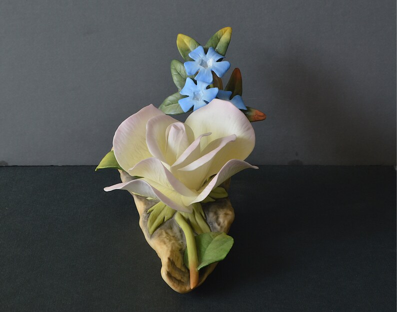 May include: A porcelain figurine of a white rose with pink highlights and blue flowers. The rose is on a brown base with green leaves.