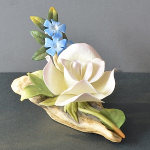 May include: A porcelain figurine of a white rose with blue forget-me-not flowers, set on a piece of driftwood.