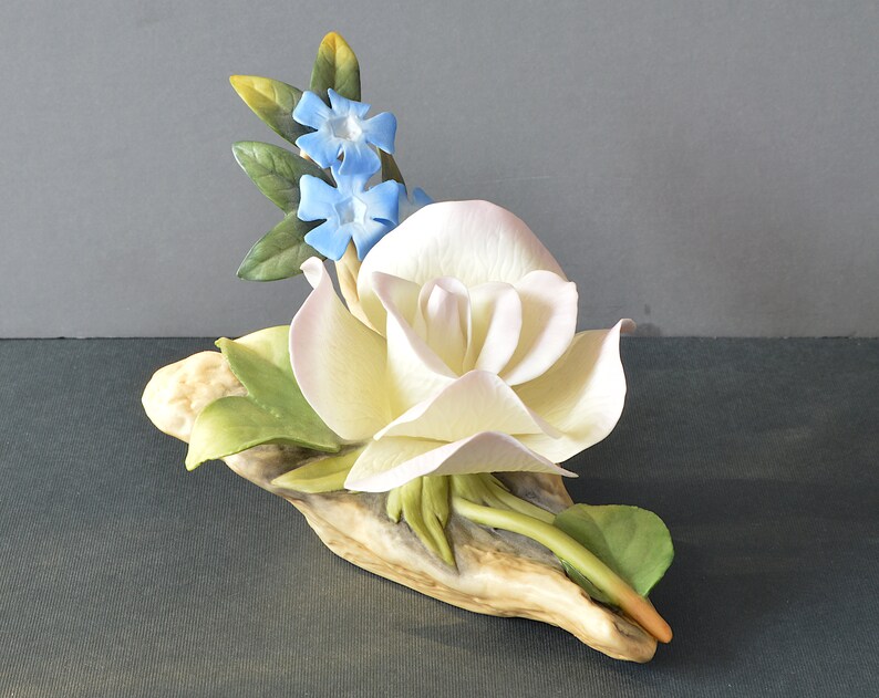 May include: A porcelain figurine of a white rose with blue forget-me-not flowers, set on a piece of driftwood.