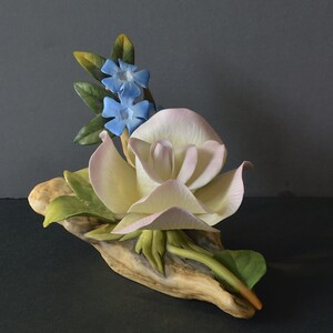 May include: A porcelain figurine of a white rose with pink edges, surrounded by green leaves and blue flowers, resting on a piece of driftwood.