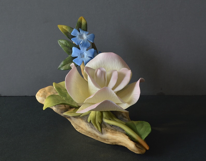 May include: A porcelain figurine of a white rose with pink edges, surrounded by green leaves and blue flowers, resting on a piece of driftwood.