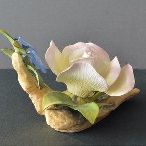 May include: A porcelain figurine of a white rose with pink edges, surrounded by green leaves and blue forget-me-not flowers.