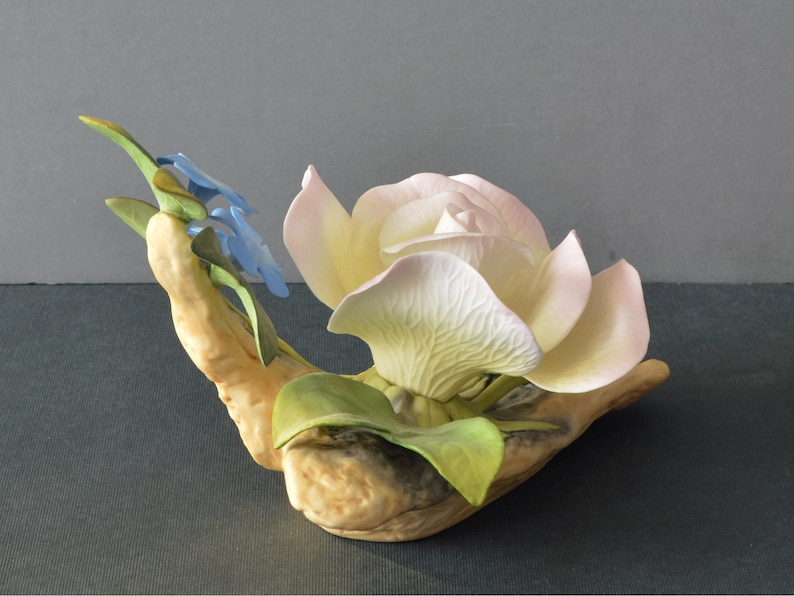 May include: A porcelain figurine of a white rose with pink edges, surrounded by green leaves and blue forget-me-not flowers.