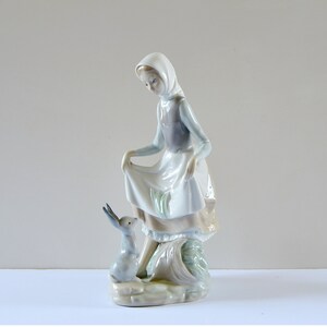 May include: A porcelain figurine of a girl in a white dress and blue hooded cloak, holding a basket and looking down at a small gray rabbit.