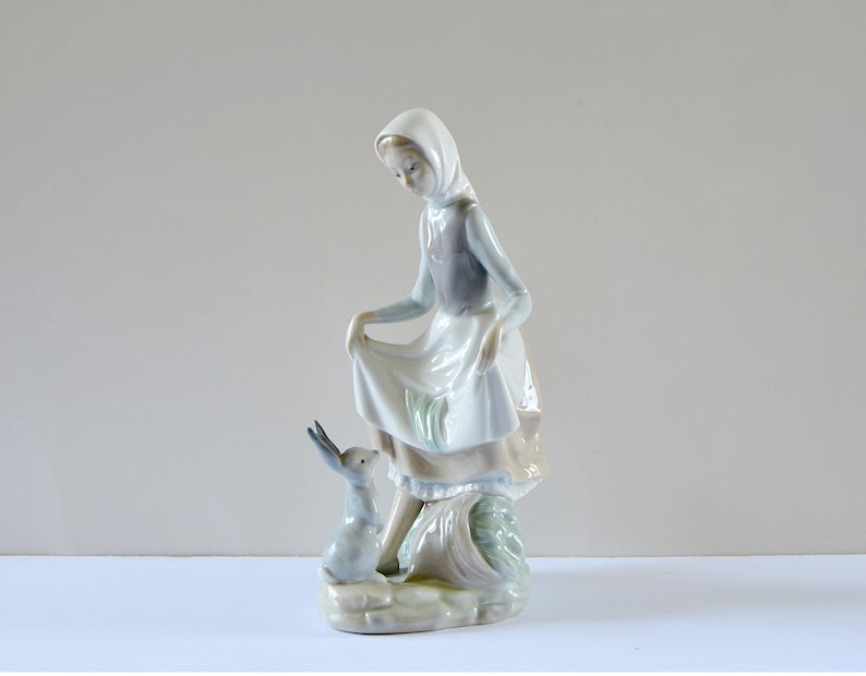 May include: A porcelain figurine of a girl in a white dress and blue hooded cloak, holding a basket and looking down at a small gray rabbit.