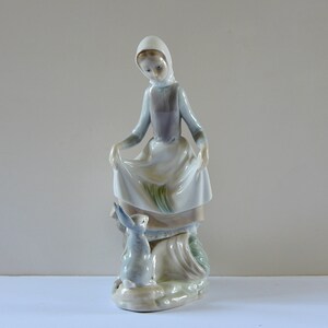 May include: A porcelain figurine of a woman in a white dress and blue hooded cloak, holding a white cloth in her hands. She is looking down at a small rabbit figurine.