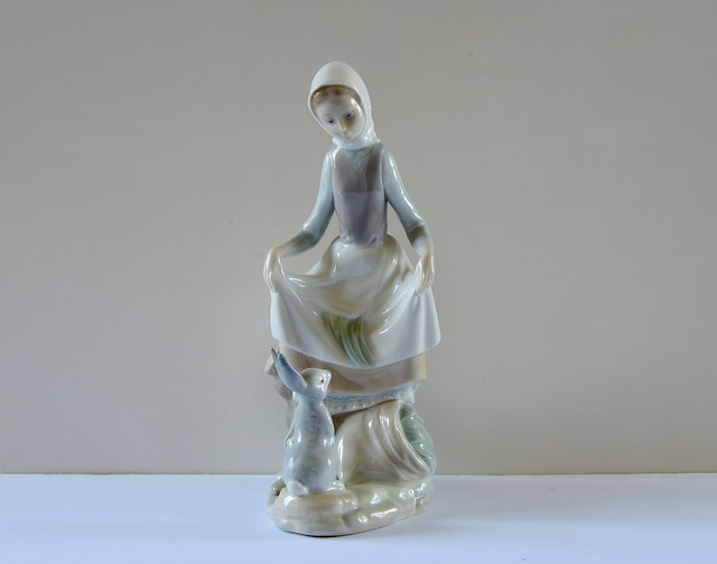 May include: A porcelain figurine of a woman in a white dress and blue hooded cloak, holding a white cloth in her hands. She is looking down at a small rabbit figurine.