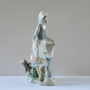 May include: A porcelain figurine of a girl in a blue and white dress and a white bonnet, holding a bouquet of flowers and standing next to a rabbit. The figurine is set on a base with green foliage.