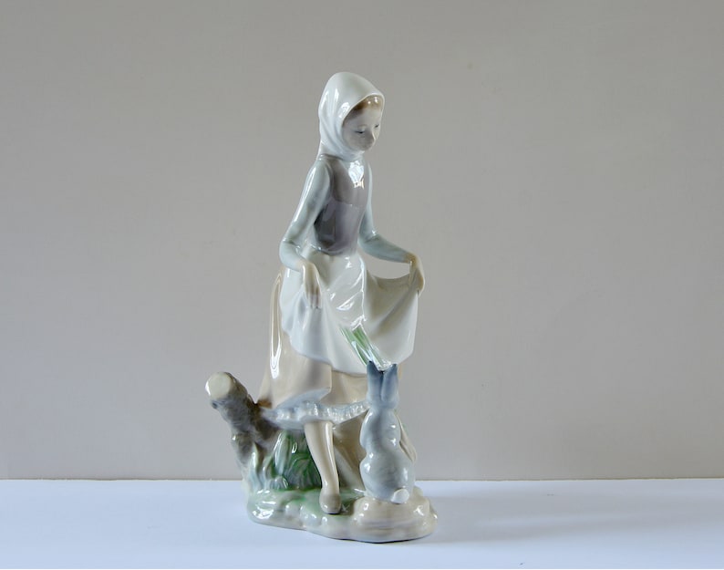 May include: A porcelain figurine of a girl in a blue and white dress and a white bonnet, holding a bouquet of flowers and standing next to a rabbit. The figurine is set on a base with green foliage.