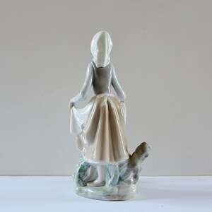 May include: A porcelain figurine of a girl in a white bonnet and brown dress, holding a basket and standing with a dog by her side.