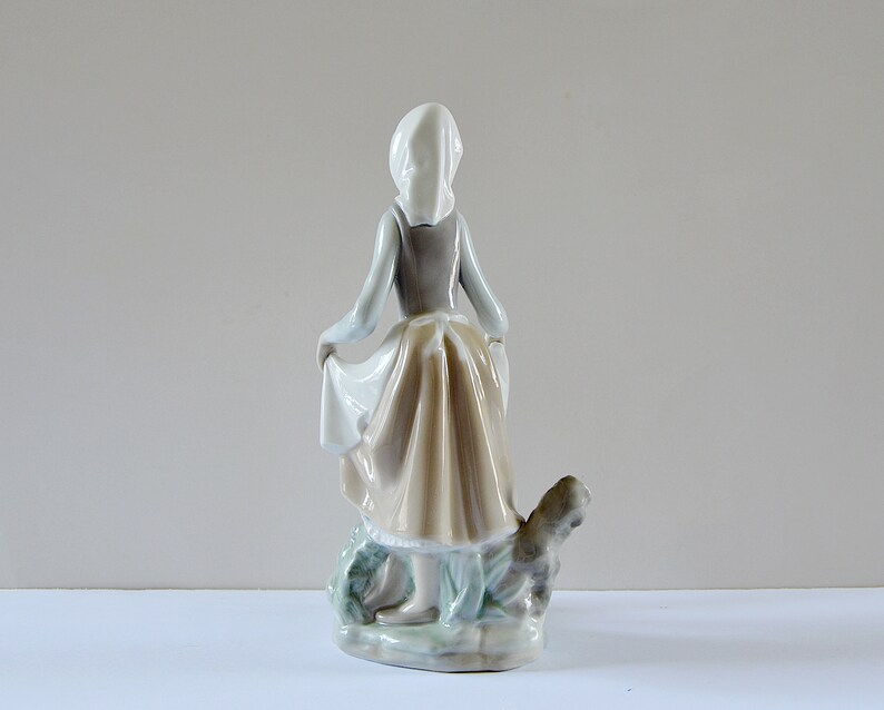 May include: A porcelain figurine of a girl in a white bonnet and brown dress, holding a basket and standing with a dog by her side.