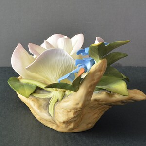 May include: A porcelain figurine of a white and pink flower with blue flowers nestled in a sculpted wooden base.