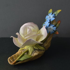 May include: A porcelain figurine of a white rose with pink edges and blue flowers, set on a brown, textured base.
