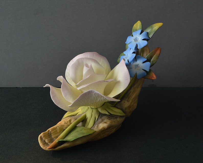 May include: A porcelain figurine of a white rose with pink edges and blue flowers, set on a brown, textured base.