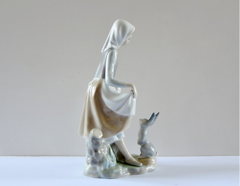May include: A porcelain figurine of a girl in a white dress and a blue hooded cloak, standing on a tree stump and looking at a small rabbit figurine.