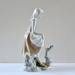 May include: A porcelain figurine of a girl in a white dress and a blue hooded cloak, standing next to a small rabbit. The girl is holding the edge of her skirt and looking down at the rabbit.
