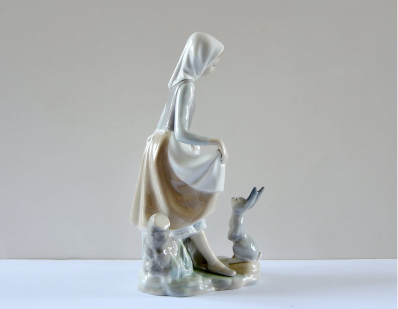 May include: A porcelain figurine of a girl in a white dress and a blue hooded cloak, standing next to a small rabbit. The girl is holding the edge of her skirt and looking down at the rabbit.