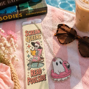 May include: A rectangular bookmark featuring a skeleton in a hat and sunglasses reading a book on a lounge chair, with the text "SCREAM QUEENS READ POOLSIDE." Attached by a silver chain is a ghost charm wearing a pink flamingo pool float.